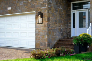 Front view of a house with a white garage door, stone exterior, and steps leading to a white front door with potted flowers, illuminated by the Urban Ambiance UQL1780 Classic Outdoor Wall Sconce in matte black from the Nicosia Collection.