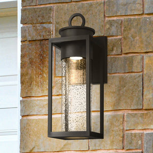 The Urban Ambiance UQL1781 Classic Outdoor Wall Sconce from the Nicosia Collection, featuring a matte black finish and marine-grade construction, is mounted on a brick wall beside a white door frame.