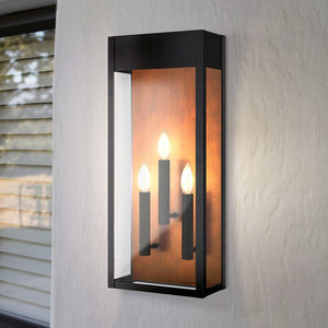 The Urban Ambiance UQL1802 Kingsley Collection Luxe Industrial Outdoor Wall Sconce features a matte black finish, marine-grade construction, glass front, and three candle-style bulbs; shown mounted on a white wall beside a window with blinds.
