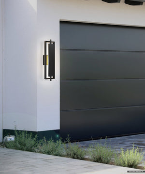 The Urban Ambiance UQL1840 Modern Outdoor Wall Sconce (20.50''H x 5.75''W), featuring a matte black finish and marine-grade build from the Juniper Collection, is mounted by a dark gray garage door on a white wall with green plants below.