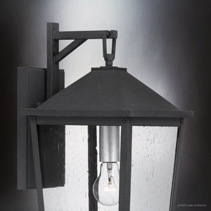 A close-up of the Urban Ambiance UQL1871 Modern Rustic Outdoor Wall Sconce from the Elmwood Collection, featuring a mottled black finish, clear glass panels, and visible bulb against a gradient backdrop. Size: 19.75