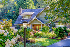 A two-story house with green siding and shingle roof is surrounded by lush landscaping, featuring the Urban Ambiance UQL1874 Modern Rustic Outdoor Post Light from the Elmwood Collection in a mottled black finish, with trees in the background.