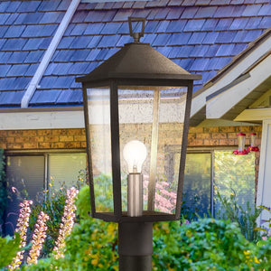 Close-up of the Urban Ambiance UQL1874 Modern Rustic Outdoor Post Light from the Elmwood Collection, featuring a mottled black finish and marine-grade construction, set in front of a shingled house and garden.