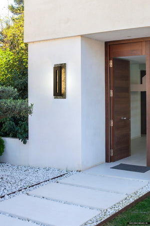Modern house entrance with a wooden door, white exterior walls, stone slab pathway bordered by white gravel, and striking architectural lighting from the Urban Ambiance UQL1880 Luxe Industrial Outdoor Wall Sconce in Earth Black.