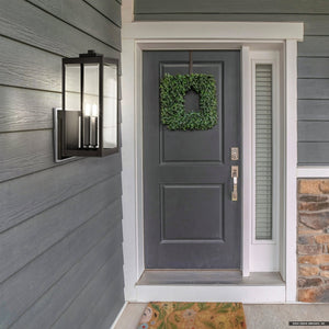A modern front door with a green wreath, adjacent narrow window, gray siding, part of a stone accent wall, and an Urban Ambiance UQL1892 Modern Farmhouse Outdoor Wall Sconce in a natural black finish from the Quincy Collection.