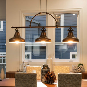 Three Sonoma Collection UQL2285 Industrial Chandeliers by Urban Ambiance, featuring industrial-inspired detailing and a Parisian Bronze finish, hang above the dining table adorned with patterned chairs and decorative vases in a room with two large windows, adding an elegant touch to the space.