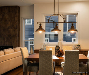Modern dining area with a wooden table, six patterned chairs, and the Urban Ambiance UQL2285 Industrial Chandelier (23.5″H x 52.5″W, Parisian Bronze Finish) from the Sonoma Collection; sofa and windows revealing a blue exterior house in the background.