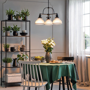 A dining area featuring a green tablecloth, a vase of white flowers, a black shelf adorned with plants and dishes, and the Urban Ambiance UQL2332 Industrial Chandelier from the Pasadena Collection (15.5″H x 24″W, Parisian Bronze Finish) suspended above the table, with natural light filtering in through sheer curtains by the window.