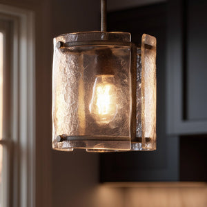 A close-up of the Urban Ambiance UQL2871 Industrial Pendant from the Nantes Collection, featuring 10-inch height and 9.25-inch width with a Silver Etch finish and textured panels, illuminated and hanging in a room with a window and dark cabinets in the background.