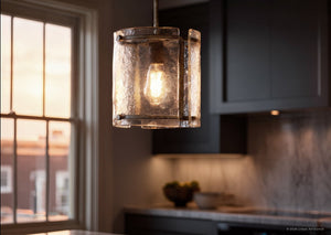 A close-up of the UQL2871 Industrial Pendant from Urban Ambiance’s Nantes Collection, featuring a textured glass shade and a silver etch finish, hanging in a kitchen with a window and cabinets in the background.