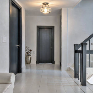 A modern hallway with light gray walls and floors, three black doors, a vase with branches, a glass-paneled staircase railing on the right, and the Urban Ambiance UQL3280 Utilitarian Ceiling Light from the Clearwater Collection (10″H x 12″W, brushed nickel finish) illuminating the space.