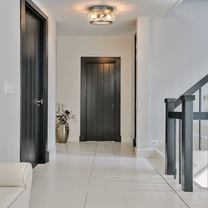 A hallway with white floors and walls, three black doors, a staircase with black railing, and a decorative vase with branches is illuminated by the Urban Ambiance UQL3290 Utilitarian Ceiling Light from the Clearwater Collection, featuring a brushed nickel finish and measuring 5.75 inches high by 12 inches wide.