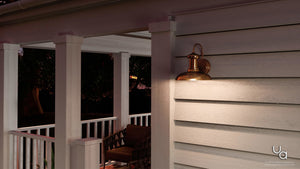 The UHP1023 Luxe Industrial Outdoor Wall Light from Urban Ambiance’s Palermo Collection, featuring a solid copper finish and measuring 10.75″ high by 10″ wide, is wall-mounted on the exterior of a white-paneled porch with a chair and railing visible, offering vintage charm and an industrial outdoor lighting style.