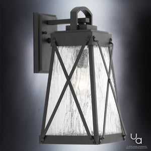 The UHP1053 English Country Outdoor Wall Light from the Saint Paul Collection by Urban Ambiance features a midnight black metal frame, clear textured glass panels, and an exposed light bulb, beautifully illuminated against a gray background. This English-inspired design adds timeless elegance and charm to any exterior space.