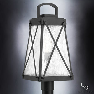 The UHP1057 English Country Outdoor Post/Pier Light from Urban Ambiance’s Saint Paul Collection, measuring 21-3/4″ high by 10-1/2″ wide in Midnight Black, features a textured glass shade, distinctive X-shaped metal accents, and a top handle. Shown illuminated against a neutral background, it evokes the timeless charm of classic vintage lanterns.