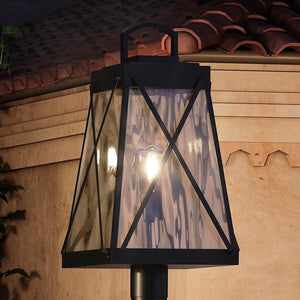 The Urban Ambiance UHP1057 English Country Outdoor Post/Pier Light from the Saint Paul Collection features a vintage lantern design with clear glass panels and a visible bulb, finished in Midnight Black. Shown mounted on a post in front of a tan stucco wall with a red tile roof in the background, this light measures 21-3/4″ high by 10-1/2″ wide.