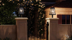 A black wrought iron gate is flanked by two stone pillars, each topped with an Urban Ambiance UHP1057 English Country Outdoor Post/Pier Light from the Saint Paul Collection, featuring a vintage lantern design in midnight black (21-3/4″H x 10-1/2″W). Flowering vines climb the gate and wall, while a stucco house with red shutters stands in the background at dusk.