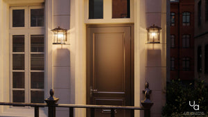 A cream-colored building at dusk features a brown front door with a window above it, flanked by two Urban Ambiance UHP1071 Rustic Outdoor Wall Lights from the Gold Coast Collection. Each coffee bronze-finished sconce measures 11.25 inches high by 8.5 inches wide and showcases a seeded glass lantern design, positioned next to a nearby window.