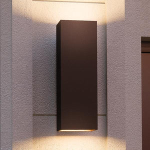 The UHP1110 Minimalist Outdoor Wall Light from the Madrid Collection by Urban Ambiance, featuring an 18″H x 6″W rectangular silhouette and an elegant Olde Bronze finish, is mounted on a textured light-colored wall and casts symmetrical up-and-down lighting in a sleek, modern style.