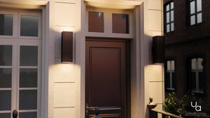 A brown front door is centered between two glass panels and flanked by Urban Ambiance UHP1110 Minimalist Outdoor Wall Lights from the Madrid Collection, each crafted with an 18-inch height and 6-inch width in an Olde Bronze finish, adding sleek and minimalist lighting to the light-colored building façade.