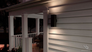 The UHP1112 Minimalist Outdoor Wall Light from Urban Ambiance’s Madrid Collection, finished in midnight black and measuring 18 inches high by 6 inches wide, is mounted on the siding of a house near a covered porch with white railings and outdoor seating at dusk. Its die-cast aluminum construction ensures both durability and style, perfectly complementing the evening ambiance.