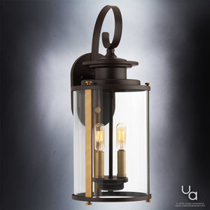 The Urban Ambiance UHP1120 Rustic Outdoor Wall Light from the Plymouth Collection features an Olde Bronze finish, a curved arm design, and a cylindrical clear glass cover with two exposed bulbs. Measuring 19.25 inches high by 8 inches wide, this modern rustic wall sconce is mounted on a wall and photographed against a neutral background.
