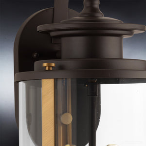 Close-up of the Urban Ambiance UHP1120 Rustic Outdoor Wall Light from the Plymouth Collection, showcasing its 19.25″ height and 8″ width in an Olde Bronze finish with clear glass cover and brass accents, mounted against a gradient background—ideal as a rustic modern wall sconce.