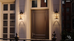 A closed brown door is centered between two Urban Ambiance UHP1120 Rustic Outdoor Wall Lights from the Plymouth Collection, featuring an Olde Bronze finish, mounted on a light-colored building facade at dusk.