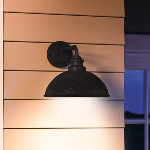 The UHP1177 Hammered Outdoor Wall Light from the Firenze Collection by Urban Ambiance, featuring a midnight black finish and measuring 10-3/8″ x 12″, is mounted on light tan horizontal siding, casting white light downward. A window frame partially appears on the right, highlighting the sconce’s distinctive industrial design and striking midnight black color.