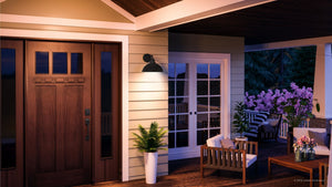At dusk, a porch with a wooden door and potted plants offers comfortable outdoor seating illuminated by the UHP1177 Hammered Outdoor Wall Light from Urban Ambiance’s Firenze Collection, featuring a 10-3/8″ x 12″ fixture in a Midnight Black finish. Beyond the door, the patio presents a table and chairs set beside blooming purple flowers, blending inviting comfort with understated industrial style.