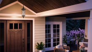 Evening view of a porch with a wooden door, glass double doors, potted plants, outdoor seating, and flowering bushes in the background, softly illuminated by the UHP1182 Rustic Outdoor Pendant Light (22″ x 7-1/2″, Olde Bronze Finish) from the Brussels Collection by Urban Ambiance.