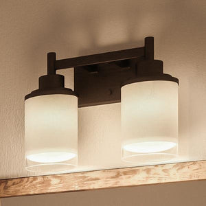 The Urban Ambiance UHP2011 Contemporary Bathroom Vanity Light from the Cupertino Collection features two cylindrical frosted glass shades and a dark Olde Bronze-finished metal base, measuring 9.5″ high by 13″ wide; mounted above a wooden trim, it provides illuminated contemporary vintage style to your space.