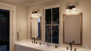 A double bathroom vanity with two mirrors is complemented by contemporary vintage lighting, including the Urban Ambiance UHP2011 Contemporary Bathroom Vanity Light from the Cupertino Collection (9.5″H x 13″W) in an Olde Bronze finish, along with black faucets, a central vase with branches, and a wall-mounted towel—all set within a neutral, modern space.