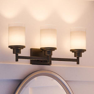 The Urban Ambiance UHP2013 Contemporary Bathroom Vanity Light from the Cupertino Collection, measuring 9.4375″H x 22″W and finished in Olde Bronze, features three evenly spaced cylindrical frosted glass shades above a mirror, casting a warm glow on light-colored walls—an ideal contemporary lighting solution for your bathroom.