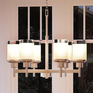 The Urban Ambiance UHP2023 Contemporary Chandelier from the Cupertino Collection, featuring a sleek brushed nickel finish and six frosted glass cylindrical shades, hangs elegantly in front of large windows at night.