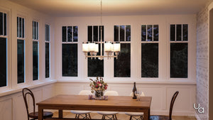 A wooden dining table with four chairs, a vase of flowers, a wine bottle, and a glass sits beneath the Urban Ambiance UHP2023 Contemporary Chandelier from the Cupertino Collection—featuring a brushed nickel finish and cylindrical shade—in a room with large windows and white walls.