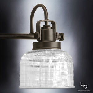 A close-up of the Urban Ambiance UHP2044 Luxe Industrial Bath Fixture from the Harlow Collection, featuring a prismatic glass shade and a curved metal arm in a Fashion Bronze finish, illuminated against a gray background.