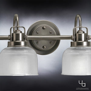 The Urban Ambiance UHP2048 Luxe Industrial Bath Fixture from the Harlow Collection, featuring an aged nickel finish and two illuminated prismatic glass shades, is shown wall-mounted against a gradient background.