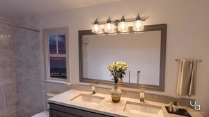 A modern bathroom featuring a spacious mirror, double sinks with a marble countertop, and wall-mounted Urban Ambiance UHP2048 Luxe Industrial Bath Fixtures from the Harlow Collection in an aged nickel finish with prismatic glass shades. A towel and a vase of white flowers adorn the vanity, while a cityscape at dusk is visible through the window.