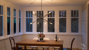 A modern dining room featuring a wooden table, four chairs, a floral centerpiece, a wine bottle, and the Urban Ambiance UHP2070 Modern Industrial Chandelier from the Miami Collection. Finished in Olde Bronze and measuring 19-5/8″ tall by 42-3/8″ wide, this striking starburst fixture adds a perfect touch of modern vintage style amid expansive windows.