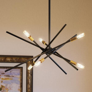 The Urban Ambiance UHP2072 Modern Industrial Chandelier from the Miami Collection, featuring an Olde Bronze finish and a striking starburst arrangement of exposed bulbs, hangs from the ceiling with part of a framed picture visible on the wall behind it.