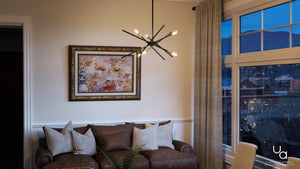 A modern living room featuring a brown leather sofa, abstract wall art, the Urban Ambiance UHP2072 Modern Industrial Chandelier from the Miami Collection in an Olde Bronze Finish, and large windows showcasing a cityscape at dusk.
