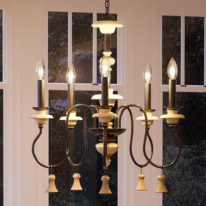 The UHP2102 French Rustic Chandelier from Urban Ambiance’s Alicante Collection, featuring a six-arm design with candle-style bulbs and an ancient bronze finish, hangs in front of large windows to add French-inspired elegance to the space.