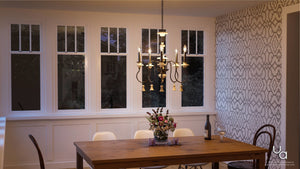Certainly! Here’s a rewritten sentence using the provided product data: A dining room featuring a wooden table, white chairs, a floral centerpiece, wine bottle and glasses, is beautifully illuminated by the Urban Ambiance UHP2102 French Rustic Chandelier from the Alicante Collection (27.875″H x 26″W, Ancient Bronze Finish); large windows and patterned wallpaper provide an elegant backdrop.