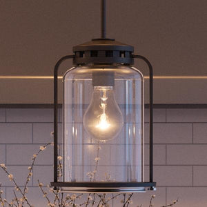 A close-up of the UHP2111 Nautical Pendant Light from the Carlsbad Collection by Urban Ambiance, featuring a lit bulb inside clear seeded glass and an Olde Bronze finish, with white subway tile and flowering branches in the background.