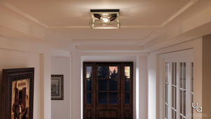 A hallway featuring the Urban Ambiance UHP2121 Luxe Industrial Flush-Mount Ceiling Fixture from the Berkeley Collection (6″H x 12″W, Charcoal Finish) adds a touch of modern rustic style overhead, accompanied by framed art on the left wall and a glass-paned wooden front door in the background illuminated by sunset.