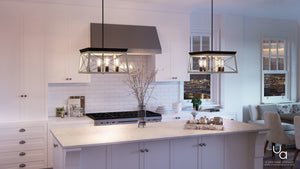 A modern kitchen features white cabinets, a marble island, and a stainless steel range hood, with the Urban Ambiance UHP2123 Luxe Industrial Chandelier (12″H x 20″W, Charcoal Finish) from the Berkeley Collection suspended above. Complementary pendant lights add to the ambiance, while a vase with branches decorates the counter and a cityscape can be seen through the window in the background.