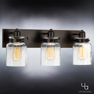 The Urban Ambiance UHP2143 Modern Farmhouse Industrial Bath Fixture from the Bridgeport Collection features three vintage-style bulbs and clear glass shades reminiscent of classic jar lighting, all mounted on a dark rectangular metal base with an elegant Olde Bronze finish—an ideal bathroom light to add timeless charm and industrial appeal to your space.