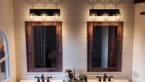 Two wooden-framed mirrors above a double sink vanity with black faucets are illuminated by the Urban Ambiance UHP2143 Modern Farmhouse Industrial Bath Fixture from the Bridgeport Collection, featuring an Olde Bronze finish and clear glass jar lighting, in a bathroom with white shiplap walls.