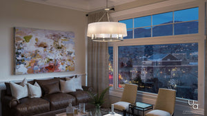 A modern living room features a brown leather sofa, abstract wall art, a glass coffee table, two chairs, and a large window framing a cityscape at dusk. The space is illuminated by the Urban Ambiance UHP2182 Cosmopolitan Chandelier from the Marsala Collection (22.375″H x 24″W) in a brushed nickel finish, which hangs gracefully overhead and brings a touch of contemporary elegance to the room.
